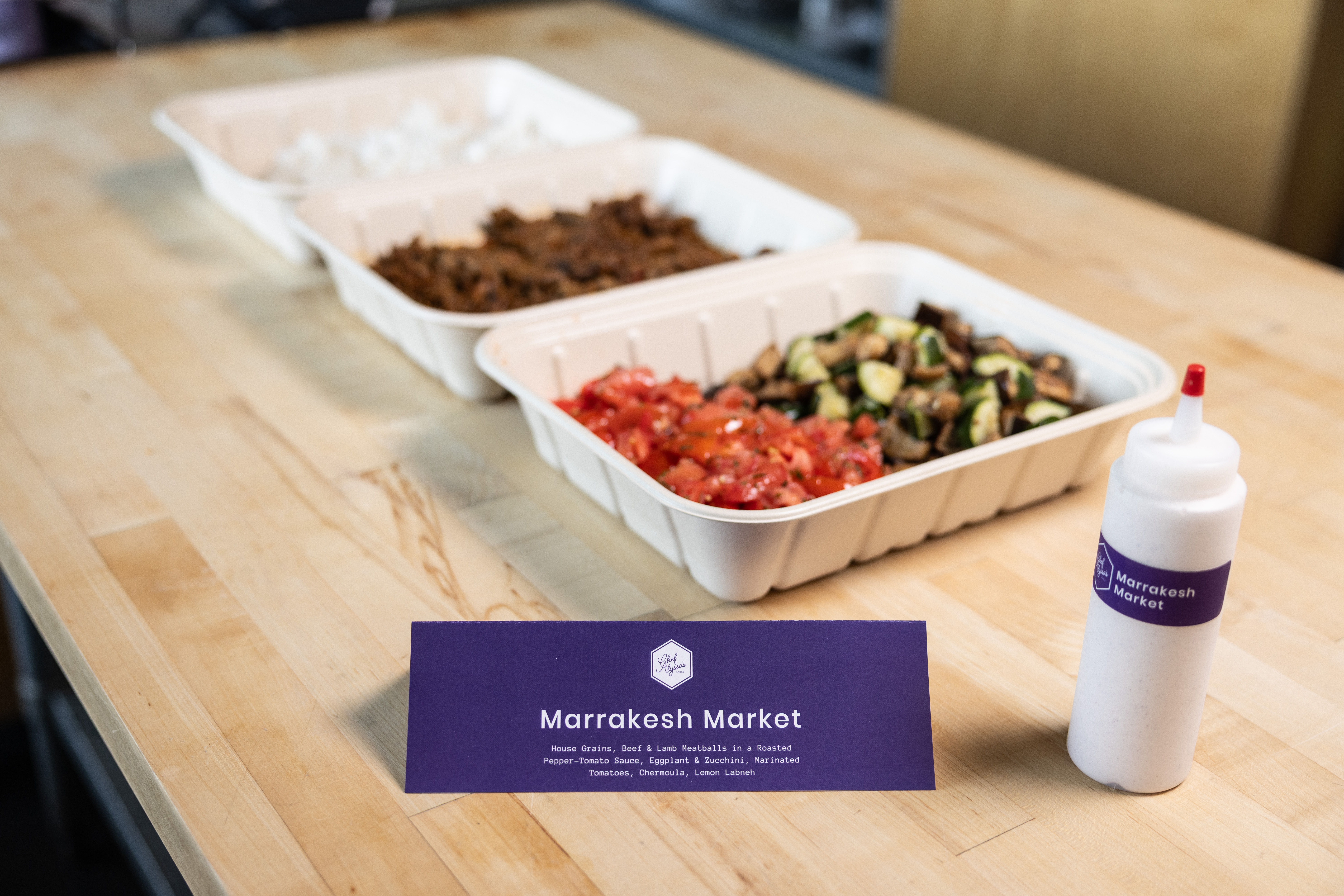 Three containers with rice, beef, and vegetables with a bottle of sauce and a placard with ingredients listed.